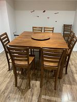Front perspective photo showing the full square wooden table with the lazy Susan centered on top and seven wooden chairs arranged around it on a wood floor.