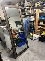 Full view of the large full-length mirror propped against a washing machine in a storage area showing the entire piece and wooden frame.