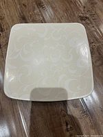 Single square decorative plate with subtle light beige floral and swirl pattern.