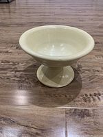 Front angle view of the off-white ceramic decorative bowl with a pedestal base on wooden floor.