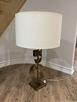 Full view of the side table lamp showing the white fabric drum shade resting on a bronze metal base with wrapped twisted metal ribbons forming a modern sculpture.
