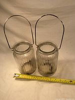 Two clear vertically ribbed glass jars with metal handles placed next to a tape measure showing size approximately 7 inches tall