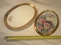 Two oval ceramic dishes with gold rims, one larger with small pink rose cluster, one smaller with large pink rose bouquet hand-painted on surface, placed next to a measuring tape showing sizes.