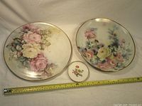 Front view of 3 decorative plates showing detailed floral patterns and gold rim. Small plate with hanger and two large plates with pastel flower designs.