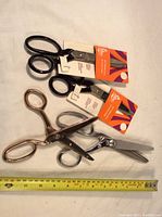 Four scissors laid flat on a brown surface next to a tape measure for length reference. Two black-handled scissors show tags indicating new condition. One silver-colored pinking shear and one copper-colored pinking shear included.