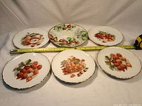 All six ceramic dessert plates arranged in a row with a measuring tape showing approximate size, placed on a plain background. Variety of fruit motif designs visible on white ceramic with gold scalloped edges.