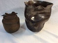 Photo showing a uniquely shaped ceramic vase beside a small ceramic lidded jar, both with dark earthy glazes, displayed on a white cloth background.