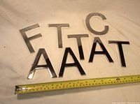 Eight stainless steel metal letters arranged on fabric, showing letters F, T, C, and several A's and T's of different sizes.