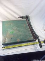 Full view of the paper cutter showing the green metal cutting surface with gridlines and long metal handle with clamp screw agreement beside a measuring tape indicating approximately 16 inches across.