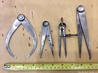 Four metallic measuring tools laid on a wooden surface with a measuring tape showing scale for size reference. Includes large and small calipers, divider compass, and large divider tool.