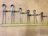 Six machinist inside calipers lined up on wood surface with measuring tape below showing size reference up to approx 17 inches.