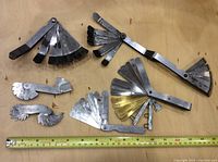 Photo showing six sets of metal machinist gauges including radius gauges and thickness gauges on a wooden surface with measuring tape for scale.