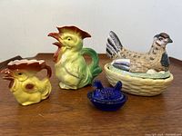 Full view of all four items: two rooster-shaped porcelain pitchers, hen on nest lidded container, cobalt glass salt cellar