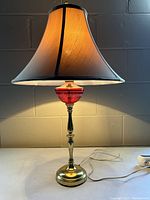 Front view of antique brass table lamp with cranberry glass bowl detail and beige fabric lampshade illuminated.