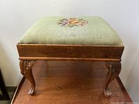 Front angle of footstool showing carved wooden legs and green needlepoint top with floral design.