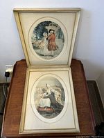 Two framed oval matted etchings showing period costume scenes, placed standing on a wooden surface