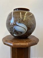 Front-side view of brown marbled swollen glass vase with white and gray swirl decor on wooden pedestal.