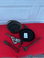 Set of cast iron pots, skillet, and kitchen tools arranged on red cloth against a white garage door.