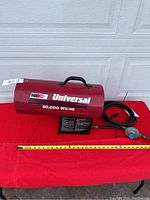 Photo of red MEC Universal propane heater with attached hose and regulator, placed against white garage door on red cloth, showing front and side view with measurement tape.