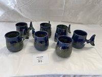 Photo showing all seven mugs in the set with their distinctive whale tail handles and blue drip glaze finish.