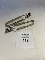 Two stainless steel escargot tongs displayed on a white surface with sale card 118