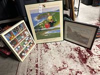 Three items on the carpet: framed 100th US Open Pebble Beach art, framed autographed golf print, and a wooden display case with golf balls and mini items.