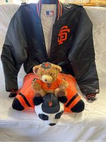 Black San Francisco Giants jacket with orange logo, stuffed animals in front including bear in Giants shirt and hat, tiger, and panda with Giants cap