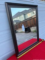 Front view of large rectangular mirror with dark wood frame against garage door outdoors. Measuring tape along bottom edge shows approximate size.