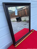 Full photo of large rectangular mirror on red surface with measuring tape showing size, lean against garage door background.