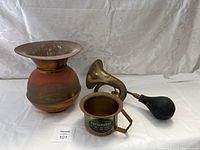 Wide shot of the three items: spittoon, bulb horn, and brass cup on white fabric background.