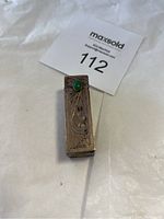 Top and side view of vintage rectangular metal lipstick holder with engraved floral pattern and green stone accent on lid, showing tarnished metal surface.