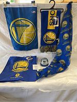 Photo showing various Golden State Warriors merchandise including scarves, flags, a tote bag, and a snow globe arranged on a white background.