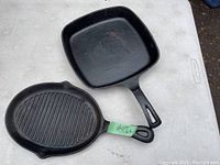 Two cast iron pans on a table, one square with a smooth surface, the other oval with ridges and both have handles.