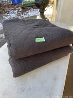 Two black folded moving blankets stacked on top of each other, showing quilted diamond stitching and rough fabric texture. Photo taken outdoors on a light stone surface.
