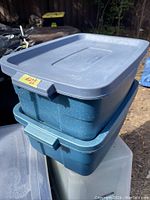 Two Rubbermaid storage bins stacked outdoors, blue color, one with speckled texture and one solid, both with lids and showing normal wear.