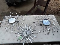 Three metal wall art pieces on table; two circular mirrors with metal arms and one sun face metal piece.