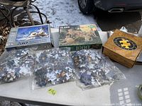 Photo showing six puzzle boxes and multiple bags of puzzle pieces, laid out on outdoor table with snow visible on ground.