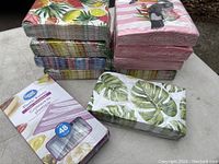 Photo showing 10 packs of colorful paper napkins with tropical and floral designs stacked together, plus a box of 48 clear plastic knives in packaging.