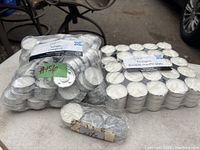 Photo showing multiple bags filled with white tealight candles in metal holders, stacked together on a concrete floor.