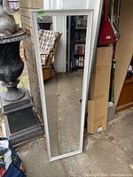 Tall rectangular wall mirror with a light-colored wood frame, set outdoors leaning against a brick wall and some stored items.