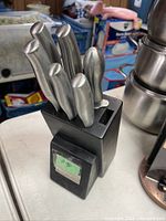 Overall view of the black knife block holding six stainless steel knives with metallic handles, on a white surface with background clutter.