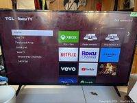 Front view of the TCL Roku flat screen TV powered on and displaying the Roku home screen interface showing various apps and streaming options.