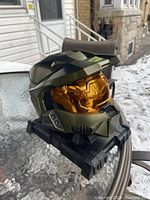 Front and right side view of green Master Chief helmet with orange visor on black display stand.
