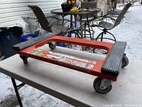 Top view of a red rectangular heavy duty dolly with black rubber grips on two corners, placed on a metal table outdoors.