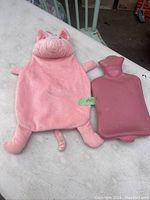 The pink rubber hot water bottle with a plush pig cover beside it, showing the cover's pig ears, snout, legs, and tail.