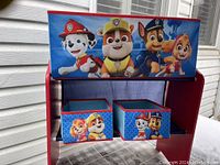 Red wooden kids shelf with three Paw Patrol themed storage bins placed inside, showing entire shelf and bins.