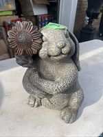 Front view of a textured gray rabbit sculpture holding a detailed metal flower with multiple petals, showing the face and flower clearly.
