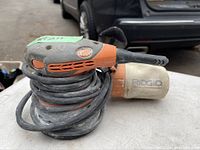 Side view of the Ridgid orbital sander showing the orange and gray design, dust collection bag, and wrapped power cord.