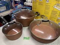 Photo showing three brown speckled cooking vessels with glass lids and black handles, arranged on a table with a yellow box in background