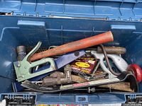 Open blue plastic tool box showing various tools including claw hammer, handsaw, metal pry bar, staple gun, staple strips and other hand tools.